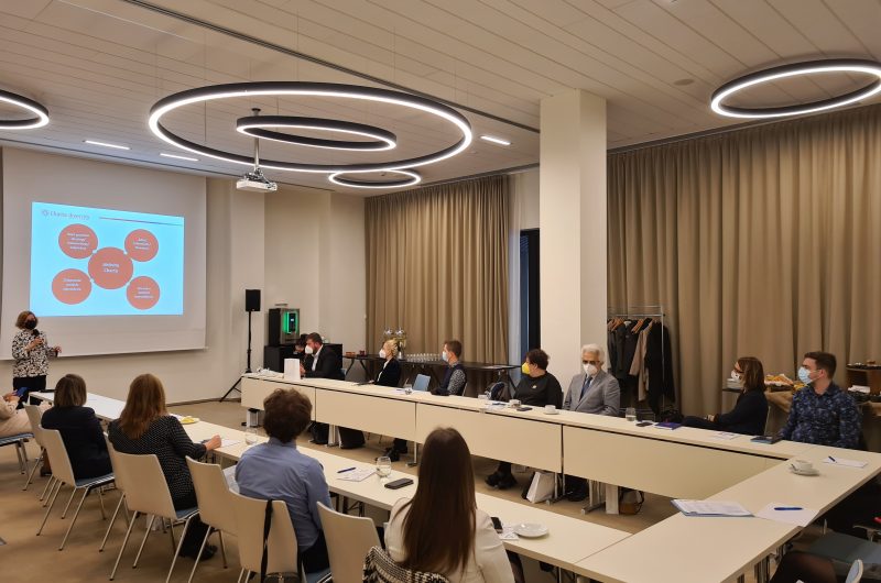 Fotka z podujatia kde ľudia sedia za stolom zatiaľ čo rečníčka hovorí do mikrofónu a prezentuje čo vidí na plátne