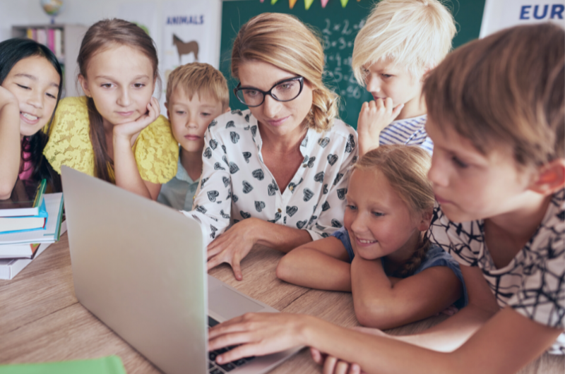 Fotobanka - Učiteľka spolu so žiakmi a žiačkami pozerajú do notebooku a usmievajú sa